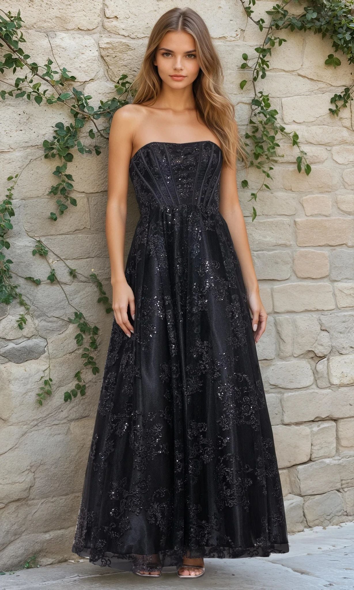 A woman stands against a stone wall, wearing the Strapless Glitter Prom Dress: Blondie Nites 4792BN in black—a fitted, floral-patterned gown with elegant sparkle and classic evening appeal.